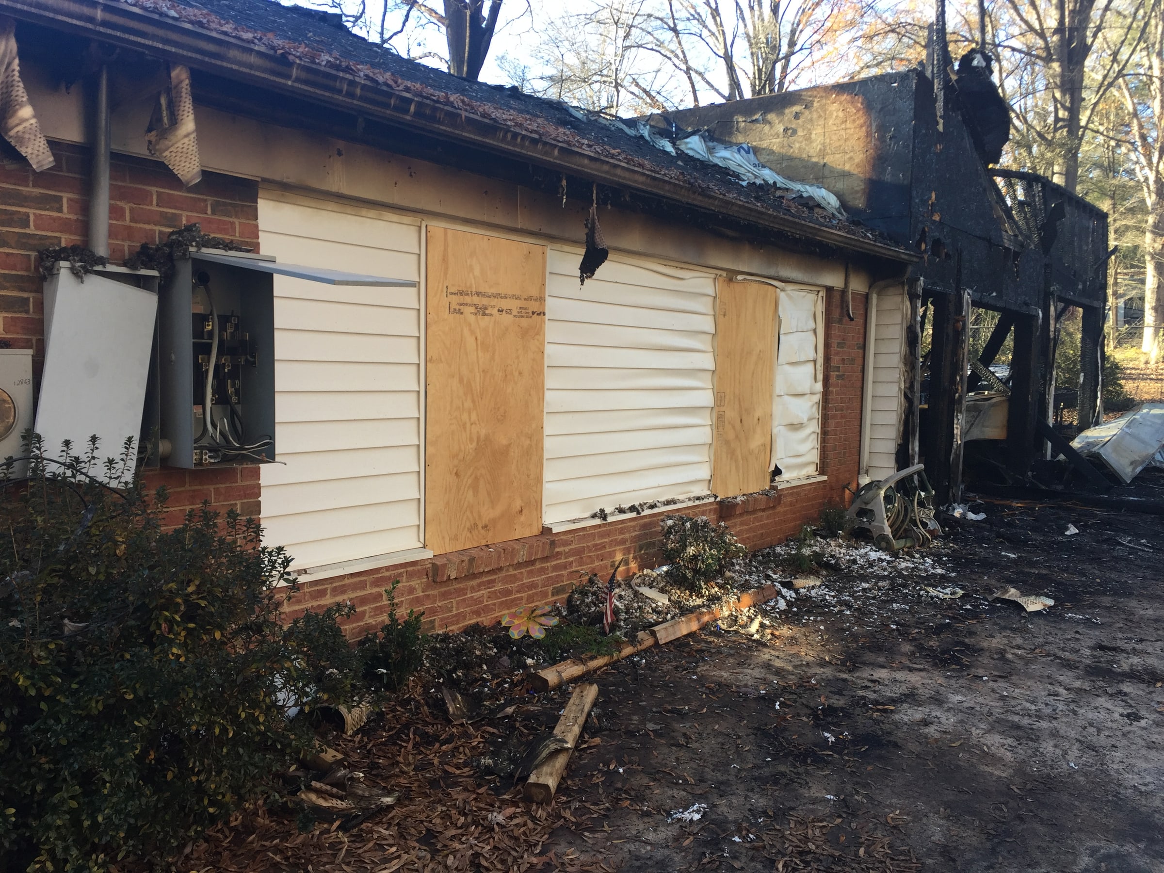 A fire-damaged single-story house seen from the street, siding charred around the windows and sheets of plywood fixed over the window openings
