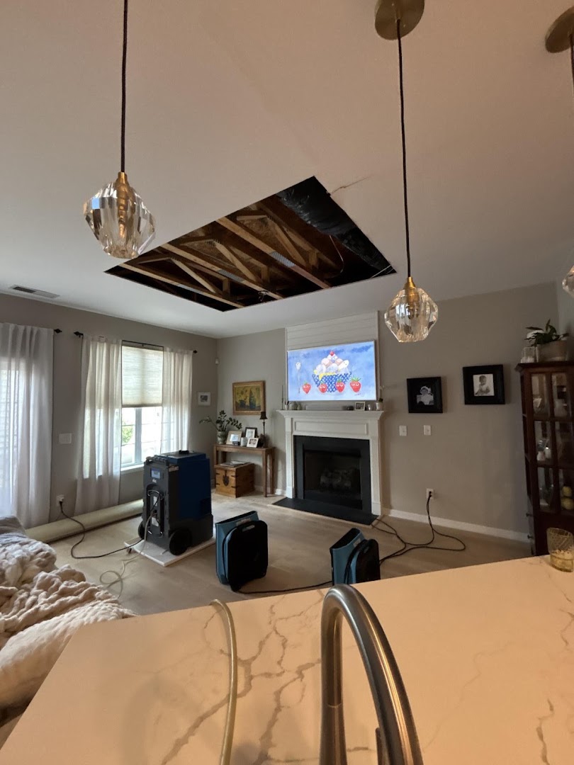 A living room with a rectangular section of ceiling cut away to the joists, a dehumidifier and air movers running below it and the room otherwise still furnished