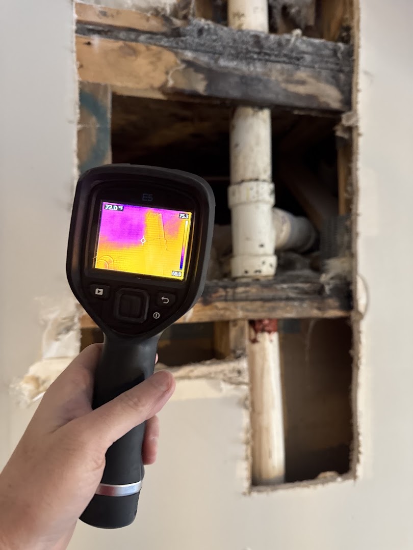 FLIR thermal camera held against an opened wall cavity with exposed studs and pipework