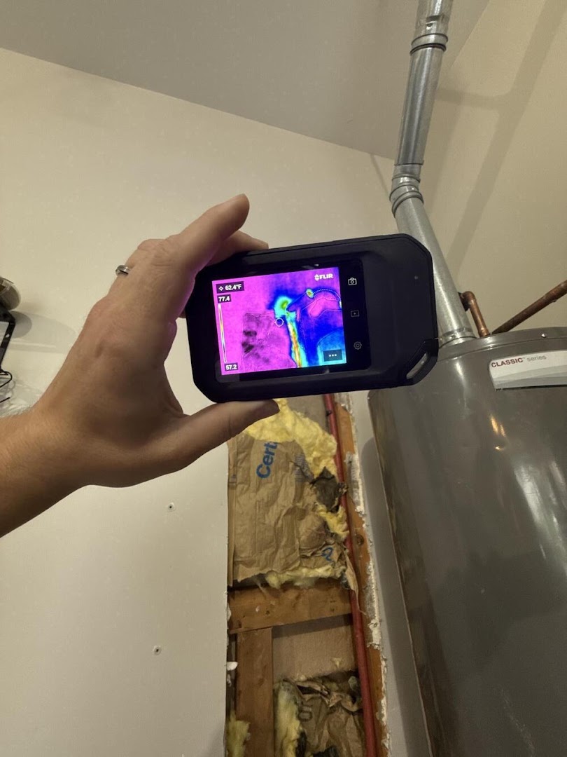A thermal imaging camera held against an opened wall cavity beside a water heater, its screen showing the cold plume where the wall is still wet