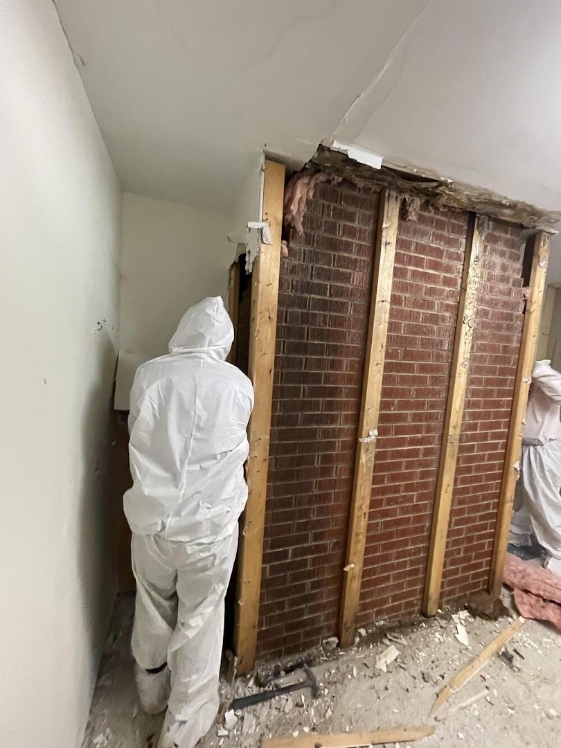 An AquaDry technician in a protective suit working beside an interior wall opened back to the brickwork